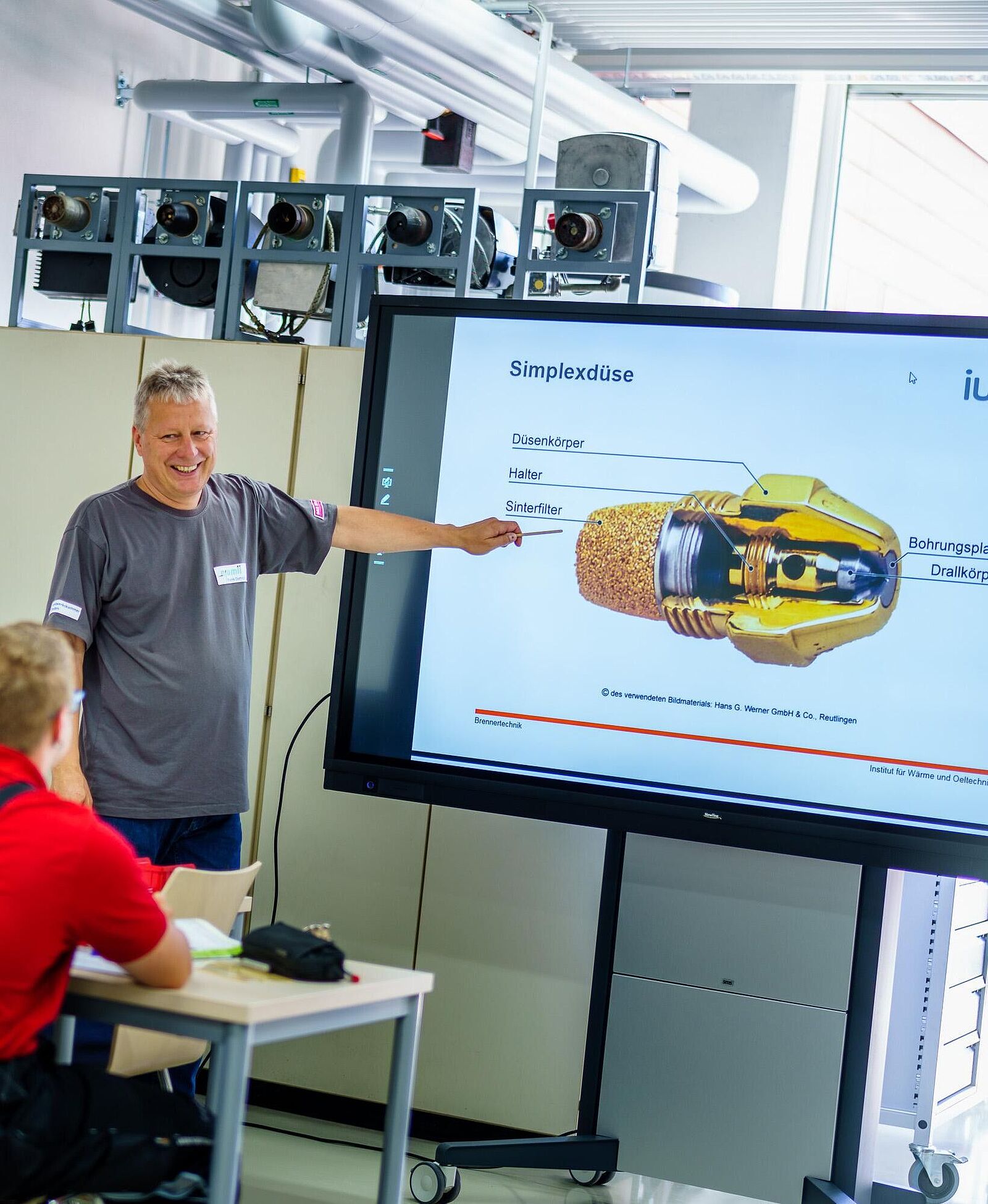 Blick in einem Unterrichtsraum der Metalltechnik. Ein Ausbilder gibt Erläuterungen anhand einer Präsentation an einer interaktiven Tafel. Zwei weitere Personen sitzen mit Unterrichtsmaterial an Unterrichtstischen.